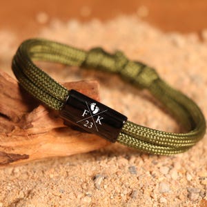 May include: Olive green paracord bracelet with a black clasp. The clasp features the initials "F K 23" with baby footprints. The bracelet is displayed on a piece of wood and sand.
