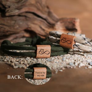 May include: Two bracelets with wooden accents featuring a heart and infinity symbol design. One bracelet is olive green, the other is beige and black. The wooden accents have the words "Forever by my side" engraved on the back.