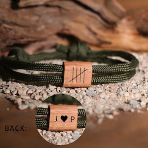 May include: A green rope bracelet with a wooden bead featuring a series of vertical lines. The back of the bead shows a heart symbol between the letters "J" and "P". The bracelet is displayed on a bed of small rocks.