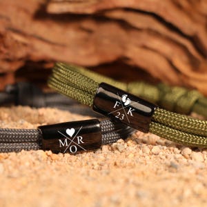 May include: Close-up of two personalized rope bracelets. One is olive green, the other is gray. Each has a black metal clasp with white text and symbols. One clasp has initials and a heart, the other has initials, the number 23, and baby footprints.