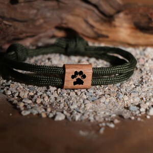 May include: An olive green rope bracelet with a wooden accent. The wooden accent features a black paw print design. The bracelet is displayed on a bed of small rocks and pebbles, with a piece of driftwood in the background.