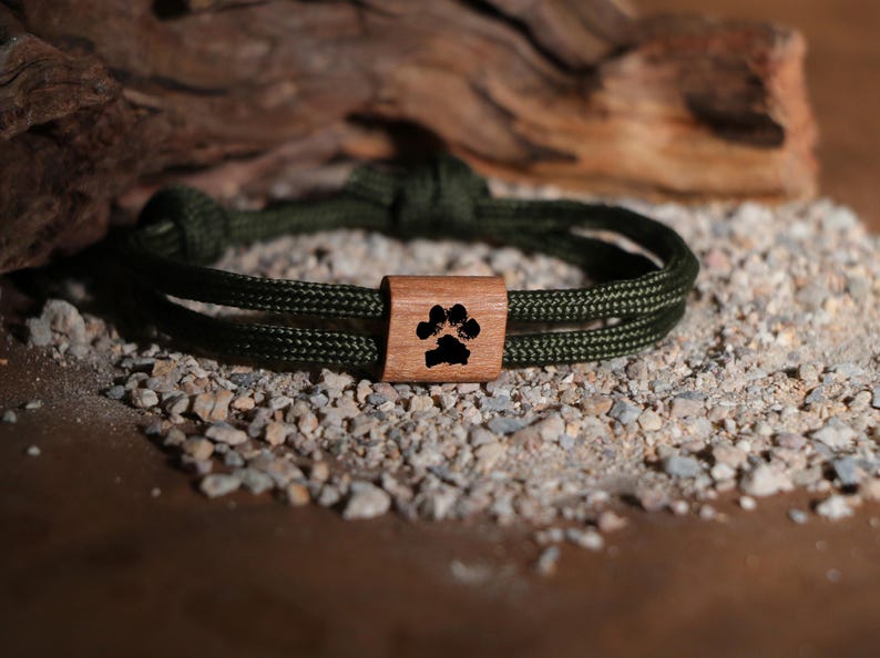 May include: An olive green rope bracelet with a wooden accent. The wooden accent features a black paw print design. The bracelet is displayed on a bed of small rocks and pebbles, with a piece of driftwood in the background.