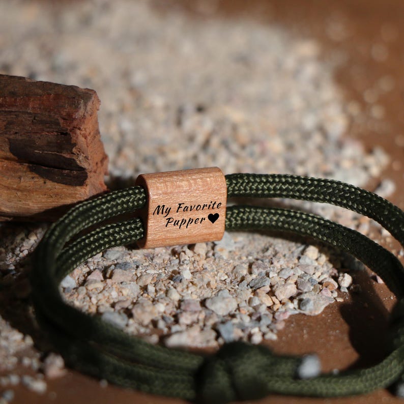 May include: A green rope bracelet with a wooden accent piece. The wooden piece is engraved with the text "My Favorite Pupper" and a heart symbol. The bracelet is displayed on a surface with small rocks and a piece of wood.