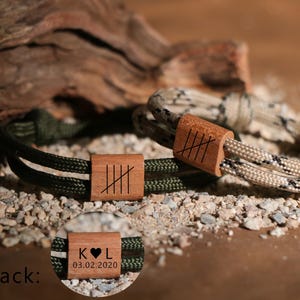 Puede incluir: Dos pulseras de paracord con detalles de madera. Una pulsera es verde oliva, la otra es beige y negra. Cada una tiene un cierre de madera con líneas grabadas. La parte posterior de una pulsera muestra iniciales y una fecha.