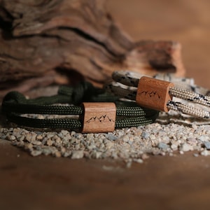 Op de afbeelding: Twee touwarmbanden met houten sluitingen met een bergontwerp. Eén armband is olijfgroen, de andere is beige en zwart. De armbanden worden gepresenteerd op kleine kiezels, met een stuk drijfhout op de achtergrond.