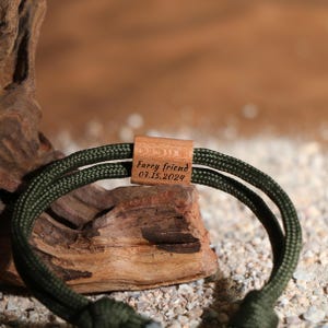 May include: A green and black rope bracelet with a wooden clasp. The wooden clasp is engraved with the words "Furry friend" and the date "07.15.2024". The bracelet is displayed on a piece of driftwood.