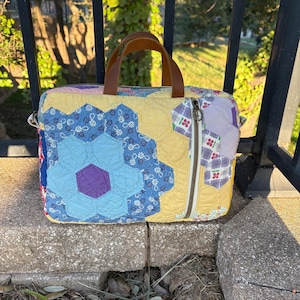 May include: A rectangular laptop bag crafted from quilted fabric featuring a patchwork of hexagonal floral patterns in shades of blue, yellow, and purple. The bag includes brown leather handles, a top zipper closure, and a vertical side zipper.