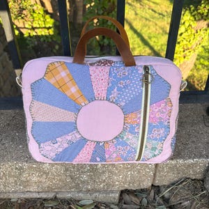 May include: A rectangular laptop bag featuring a pink fabric base with a central fan-shaped patchwork design in various floral and plaid patterns. It includes two brown leather handles, a top zipper closure, and a vertical side zipper.