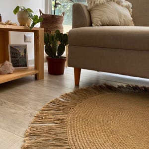 K&ouml;nnte beinhalten: Ein runder, gewebter Jute-Teppich mit Fransen. Der Teppich hat eine warme, beige Farbe und liegt auf einem hellen Holzboden. Das Wort "LEMONT" ist wei&szlig; auf dem Teppich aufgedruckt. Der Teppich befindet sich in der N&auml;he eines beigen Sofas.