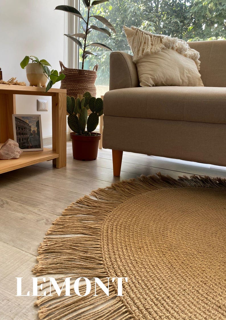K&ouml;nnte beinhalten: Ein runder, gewebter Jute-Teppich mit Fransen. Der Teppich hat eine warme, beige Farbe und liegt auf einem hellen Holzboden. Das Wort "LEMONT" ist wei&szlig; auf dem Teppich aufgedruckt. Der Teppich befindet sich in der N&auml;he eines beigen Sofas.