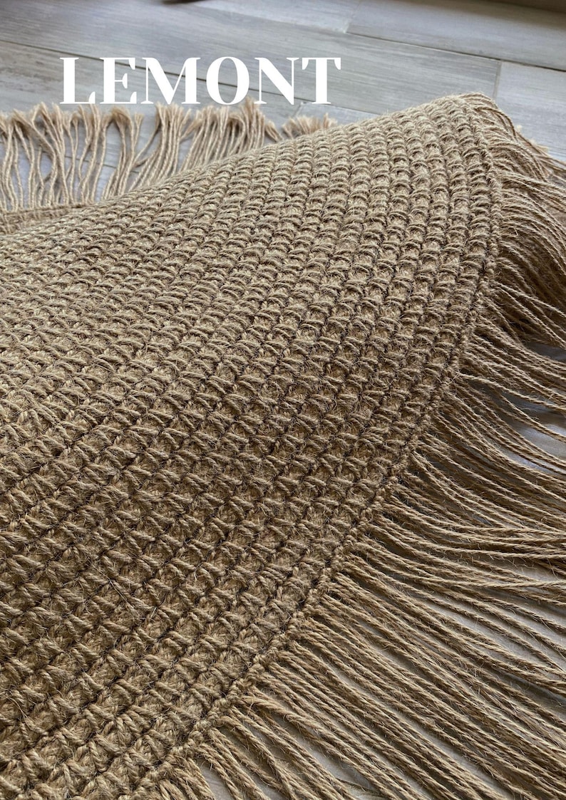 K&ouml;nnte beinhalten: Ein runder, gewebter Jute-Teppich mit strukturiertem Muster und Fransen. Der Teppich hat eine nat&uuml;rliche beige Farbe und tr&auml;gt oben das Wort "LEMONT" in wei&szlig;en Blockbuchstaben. Geeignet f&uuml;r die Inneneinrichtung.