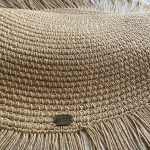 K&ouml;nnte beinhalten: Ein runder, gewebter Jute-Teppich mit Fransen. Der Teppich hat eine nat&uuml;rliche beige Farbe und zeigt oben das Wort "LEMONT" in wei&szlig;er Schrift. Ein kleines, rechteckiges Etikett mit dem Wort "Lemont" ist auf den Teppich gen&auml;ht.