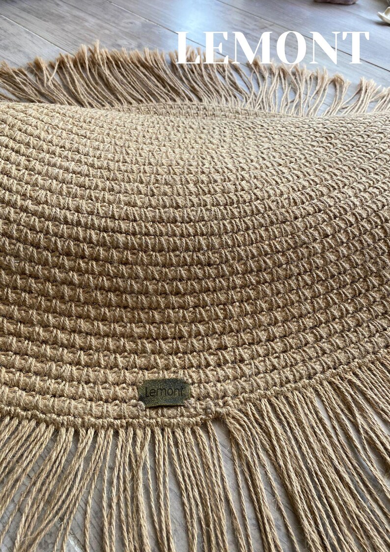K&ouml;nnte beinhalten: Ein runder, gewebter Jute-Teppich mit Fransen. Der Teppich hat eine nat&uuml;rliche beige Farbe und zeigt oben das Wort "LEMONT" in wei&szlig;er Schrift. Ein kleines, rechteckiges Etikett mit dem Wort "Lemont" ist auf den Teppich gen&auml;ht.