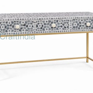 May include: A decorative console table with a geometric patterned top in shades of gray and white. The table has three drawers with white knobs and a gold-colored metal frame. The table is a stylish piece of furniture.