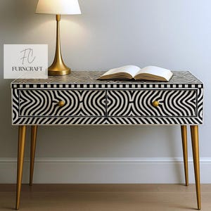 May include: A black and white patterned console table with two drawers and gold-colored legs. A gold lamp and an open book sit on top of the table. The table has the text "FURNCRAFT".