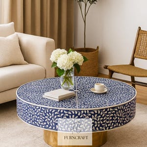 May include: A circular coffee table featuring a blue and white floral design on top and a gold-coloured base. The table is in a living room setting. A vase of white flowers and a cup and saucer are on the table. The table's diameter is approximately 90 cm.
