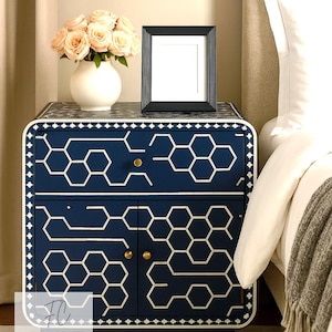 May include: A navy blue bedside cabinet with a white geometric design, gold handles, and a black picture frame. A white vase with pink roses sits on top. The cabinet is next to a bed with white bedding.
