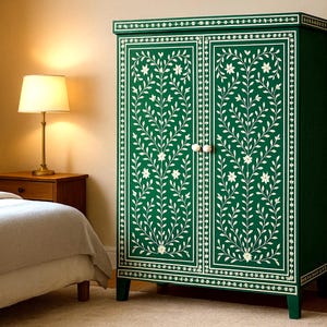 May include: A dark green cabinet with white floral and leaf patterns. The cabinet has two doors with white knobs and is decorated with a white border. The cabinet is in a bedroom setting with a lamp and a bed.