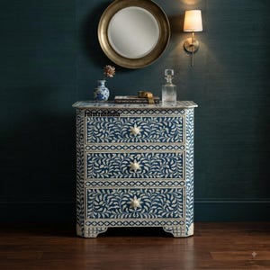 May include: A three-drawer chest featuring a deep blue finish with intricate white floral vine inlay patterns. Each drawer is equipped with a white flower-shaped knob. The piece stands on a flat base and displays a repeating geometric border design.