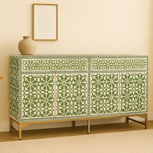May include: A green and white patterned cabinet with four doors and two drawers. The cabinet has a gold-colored base and legs. A beige vase and a framed picture are on the wall above the cabinet.