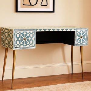 Può includere: Una scrivania decorativa con un motivo geometrico blu e bianco. La scrivania ha due cassetti con pomelli color oro e gambe sottili color oro. La scrivania è in legno e ha un interno nero.