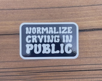 Könnte beinhalten: Ein rechteckiger Aufkleber mit abgerundeten Ecken. Der Aufkleber hat einen schwarzen Hintergrund mit weißem Text, der "NORMALIZE CRYING IN PUBLIC" lautet. Der Text ist in einer fetten Retro-Schriftart, und der Aufkleber hat einen weißen Rand.