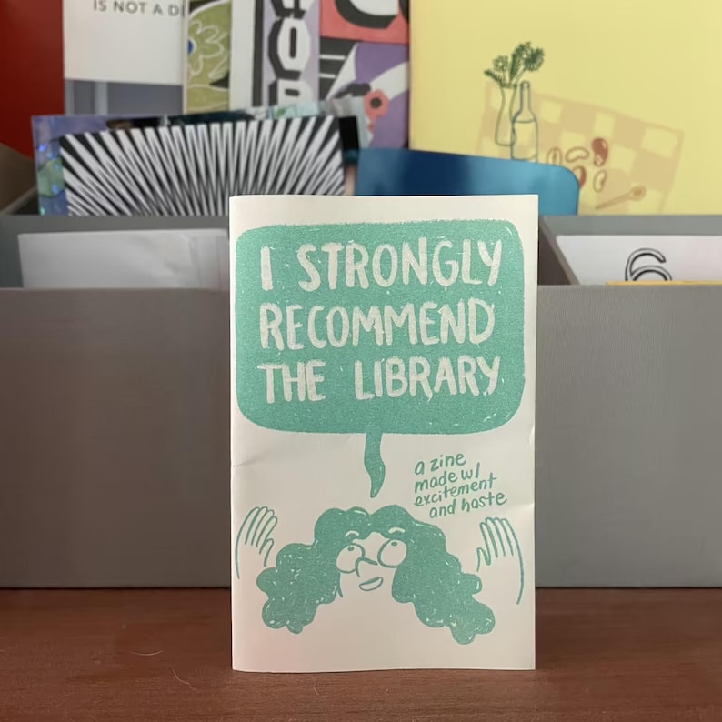 May include: A white zine with the text "I STRONGLY RECOMMEND THE LIBRARY" in a teal speech bubble. Below the bubble is a drawing of a person with teal hair and glasses. The zine also says "a zine made w/ excitement and haste."