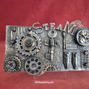 May include: Steampunk-style artwork featuring the word "STEAM" in metallic lettering. The piece incorporates gears, keys, and industrial components in silver, bronze, and gold hues, set against a textured backdrop.