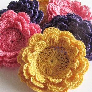 May include: Four crocheted flowers in shades of pink, purple, and yellow. The flowers are layered on top of each other, with the pink flower on top.