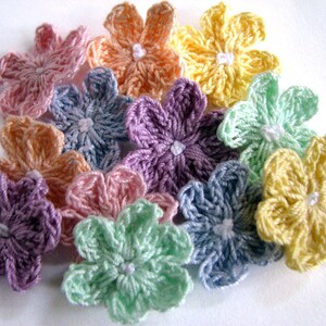 May include: A collection of ten crocheted flowers in various pastel colors, including lavender, light blue, pink, green, and yellow. The flowers are all the same size and have a five-petal design.
