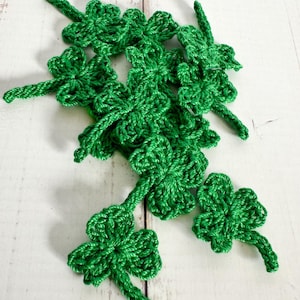 May include: A collection of handmade, bright green crochet shamrocks, each with three leaves and a stem. The shamrocks are scattered on a white wooden surface, perfect for St. Patrick's Day decorations or craft projects.