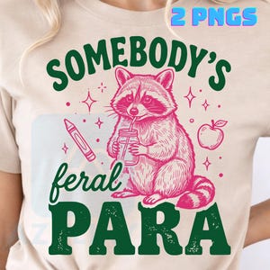 Könnte beinhalten: Ein beiges T-Shirt mit einer Grafik eines Waschbären, der ein Einmachglas mit Strohhalm, einen Buntstift und einen Apfel hält. Der Text lautet "SOMEBODY'S feral PARA" in Grün und Pink. Das Design enthält kleine Sternakzente und "2 PNGS".