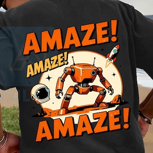 May include: A charcoal gray t-shirt featuring a graphic print of an orange robot standing on a planet surface. The design includes an astronaut helmet, a small rocket, and the text AMAZE! in large orange letters above and below the robot.