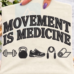 Könnte beinhalten: Ein cremefarbenes T-Shirt mit dem schwarz gestickten Text MOVEMENT IS MEDICINE in einer fetten, geschwungenen Schrift. Unter dem Text befinden sich schwarz gestickte Symbole einer Hantel, einer Kettlebell, eines Laufschuhs, eines Springseils und einer zusammengerollten Yogamatte.