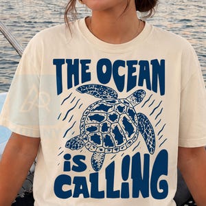 K&ouml;nnte beinhalten: Ein cremefarbenes T-Shirt mit kurzen &Auml;rmeln und einer dunkelblauen Grafik einer schwimmenden Meeresschildkr&ouml;te. Der Text "THE OCEAN is CALLING" ist in einer fetten, wellenf&ouml;rmigen dunkelblauen Schrift &uuml;ber und unter der Schildkr&ouml;te gedruckt.