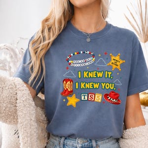 K&ouml;nnte beinhalten: Ein blaues T-Shirt mit einem Grafikdruck, der ein Perlenarmband mit der Aufschrift You've got a friend in me, einen gelben Sheriffstern, rote Cowboystiefel, einen roten Cowboyhut, TS5-Bl&ouml;cke und den Text I KNEW IT, I KNEW YOU zeigt.