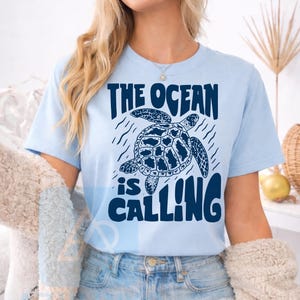 K&ouml;nnte beinhalten: Ein hellblaues T-Shirt mit kurzen &Auml;rmeln und einer dunkelblauen Grafik einer schwimmenden Meeresschildkr&ouml;te. Der Text "THE OCEAN is CALLING" ist in einer fetten, dunkelblauen Schrift &uuml;ber und unter der Schildkr&ouml;tenillustration aufgedruckt.