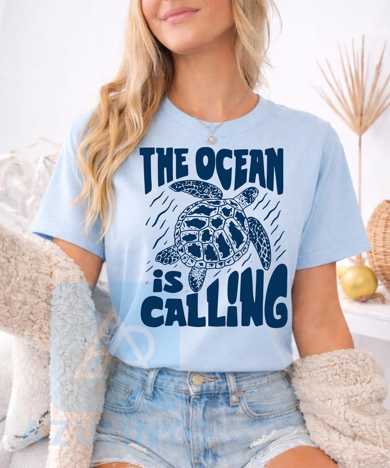 K&ouml;nnte beinhalten: Ein hellblaues T-Shirt mit kurzen &Auml;rmeln und einer dunkelblauen Grafik einer schwimmenden Meeresschildkr&ouml;te. Der Text "THE OCEAN is CALLING" ist in einer fetten, dunkelblauen Schrift &uuml;ber und unter der Schildkr&ouml;tenillustration aufgedruckt.