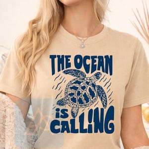 K&ouml;nnte beinhalten: Ein beiges Kurzarm-T-Shirt mit einer dunkelblauen Grafik einer schwimmenden Meeresschildkr&ouml;te. Der Text "THE OCEAN is CALLING" ist in dunkelblauen Buchstaben &uuml;ber und unter der Schildkr&ouml;tenillustration gedruckt, umgeben von wellenf&ouml;rmigen Linien.