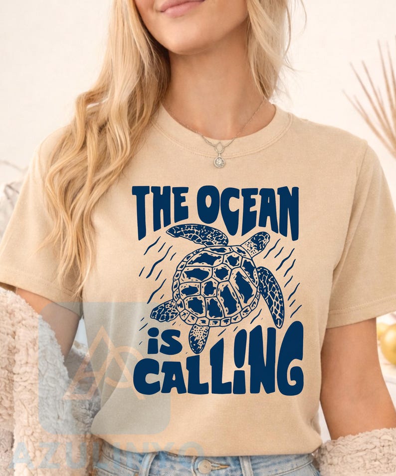 K&ouml;nnte beinhalten: Ein beiges Kurzarm-T-Shirt mit einer dunkelblauen Grafik einer schwimmenden Meeresschildkr&ouml;te. Der Text "THE OCEAN is CALLING" ist in dunkelblauen Buchstaben &uuml;ber und unter der Schildkr&ouml;tenillustration gedruckt, umgeben von wellenf&ouml;rmigen Linien.