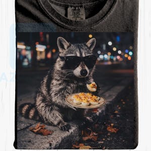 May include: A dark charcoal gray t-shirt with a Comfort Colors label at the neck. The graphic print features a raccoon wearing black sunglasses, sitting on a curb at night, holding a small plate of deviled eggs and eating one piece.