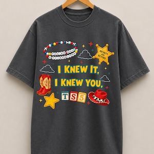 K&ouml;nnte beinhalten: Ein anthrazitfarbenes Kurzarm-T-Shirt mit einem Grafikdruck, der ein Perlenarmband, einen gelben Sheriffstern, rote Cowboystiefel, einen roten Cowboyhut und Buchstabenw&uuml;rfel mit der Aufschrift TS5 zeigt. Der Text lautet I KNEW IT, I KNEW YOU.