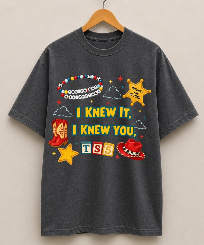 K&ouml;nnte beinhalten: Ein anthrazitfarbenes Kurzarm-T-Shirt mit einem Grafikdruck, der ein Perlenarmband, einen gelben Sheriffstern, rote Cowboystiefel, einen roten Cowboyhut und Buchstabenw&uuml;rfel mit der Aufschrift TS5 zeigt. Der Text lautet I KNEW IT, I KNEW YOU.
