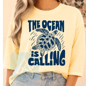 K&ouml;nnte beinhalten: Ein hellgelbes T-Shirt mit kurzen &Auml;rmeln und einer dunkelblauen Grafik einer Meeresschildkr&ouml;te auf der Brust. Der Text &uuml;ber und unter der Schildkr&ouml;te lautet THE OCEAN is CALLING in einer fetten, dunkelblauen Schrift. Rundhalsausschnitt.