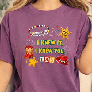 K&ouml;nnte beinhalten: Ein violettes T-Shirt mit einem Grafikdruck mit dem Text I KNEW IT, I KNEW YOU, TS5, INFINITY AND BEYOND und You've got a friend in me. Das Design enth&auml;lt ein Perlenarmband, einen gelben Sheriffstern, rote Cowboystiefel und einen roten Hut.