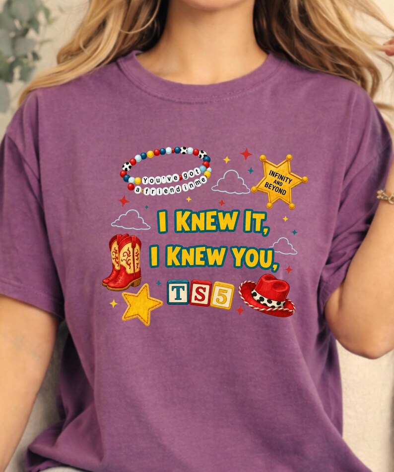 K&ouml;nnte beinhalten: Ein violettes T-Shirt mit einem Grafikdruck mit dem Text I KNEW IT, I KNEW YOU, TS5, INFINITY AND BEYOND und You've got a friend in me. Das Design enth&auml;lt ein Perlenarmband, einen gelben Sheriffstern, rote Cowboystiefel und einen roten Hut.