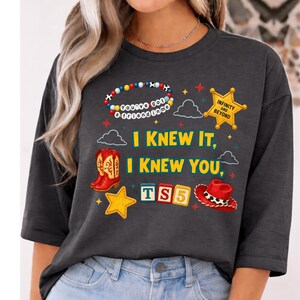 K&ouml;nnte beinhalten: Ein anthrazitfarbenes T-Shirt mit einem Grafikdruck, der ein Perlenarmband mit der Aufschrift "You've got a friend in me", einen gelben Sheriffstern mit "INFINITY AND BEYOND", rote Cowboystiefel, einen roten Hut, Buchstabenw&uuml;rfel mit "TS5" und Sterne zeigt.