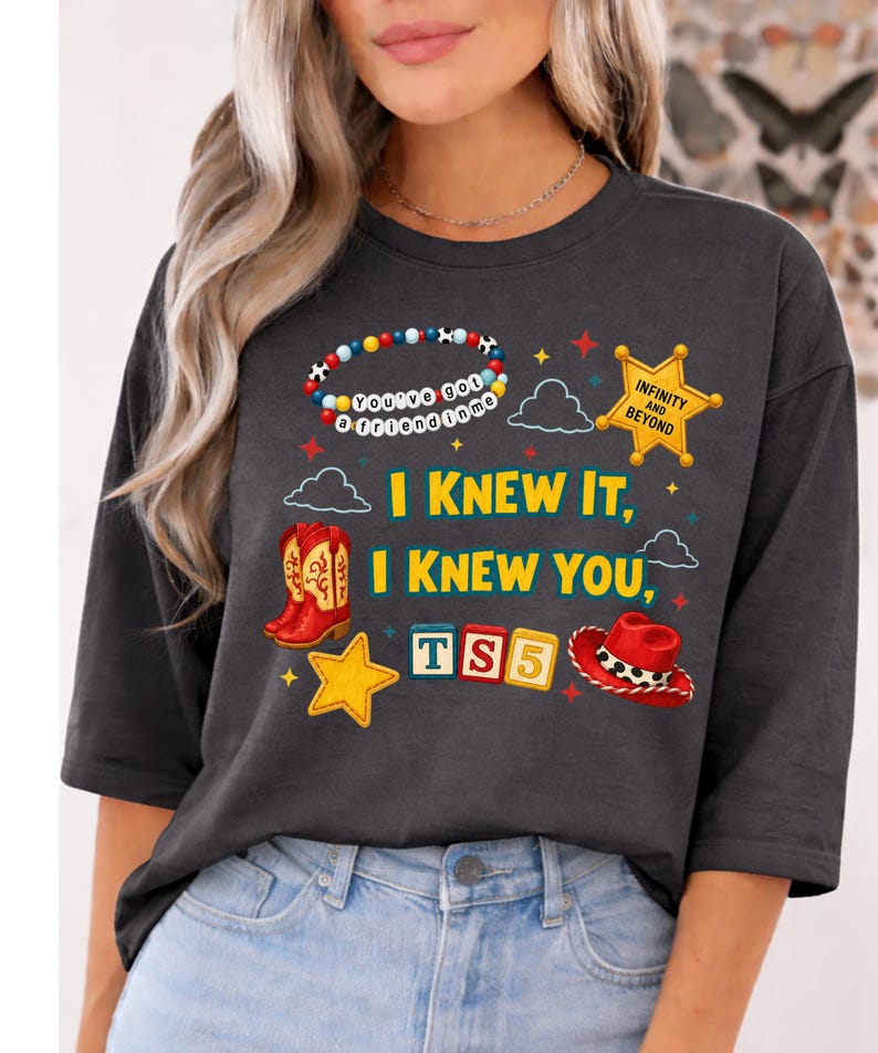 K&ouml;nnte beinhalten: Ein anthrazitfarbenes T-Shirt mit einem Grafikdruck, der ein Perlenarmband mit der Aufschrift "You've got a friend in me", einen gelben Sheriffstern mit "INFINITY AND BEYOND", rote Cowboystiefel, einen roten Hut, Buchstabenw&uuml;rfel mit "TS5" und Sterne zeigt.