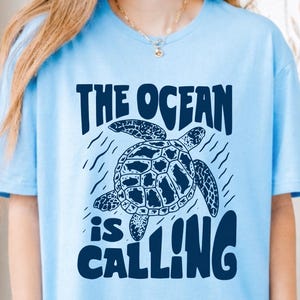 K&ouml;nnte beinhalten: Ein hellblaues T-Shirt mit einer dunkelblauen Grafik einer schwimmenden Meeresschildkr&ouml;te. &Uuml;ber der Schildkr&ouml;te steht der Text THE OCEAN und darunter der Text is CALLING. Das Design enth&auml;lt kleine wellenf&ouml;rmige Linien um die Schildkr&ouml;te.