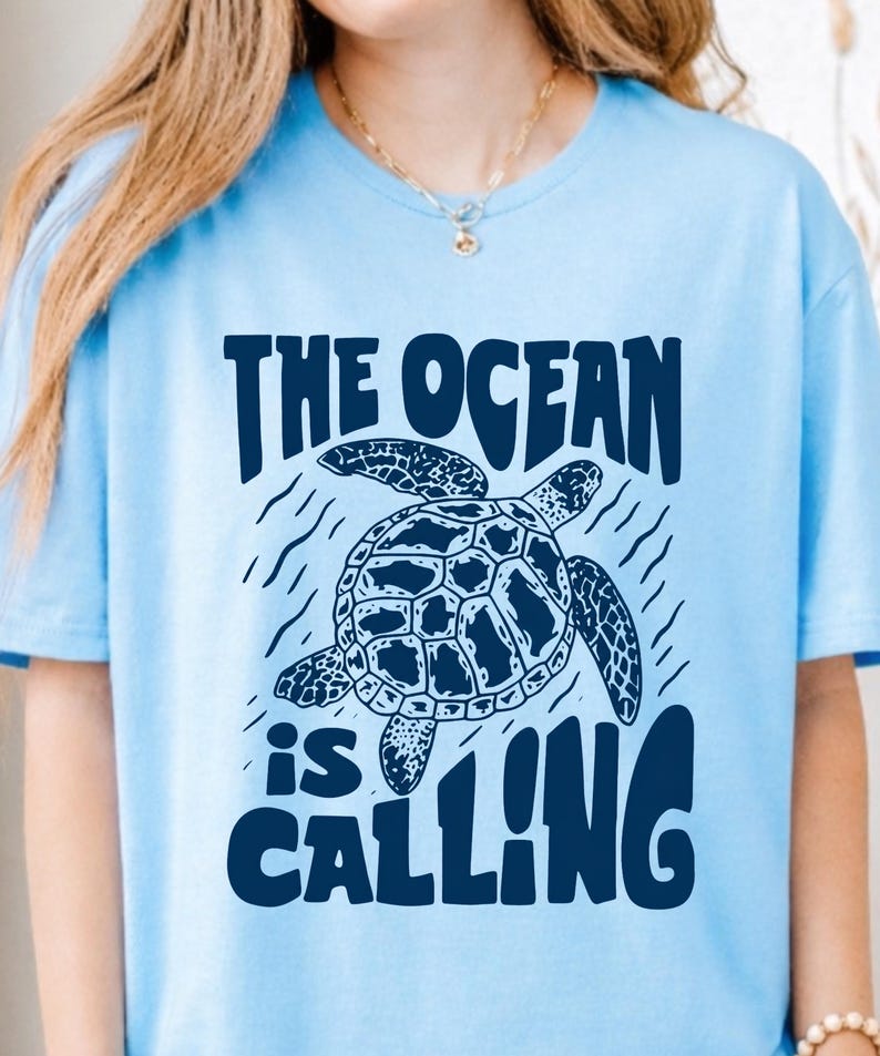 K&ouml;nnte beinhalten: Ein hellblaues T-Shirt mit einer dunkelblauen Grafik einer schwimmenden Meeresschildkr&ouml;te. &Uuml;ber der Schildkr&ouml;te steht der Text THE OCEAN und darunter der Text is CALLING. Das Design enth&auml;lt kleine wellenf&ouml;rmige Linien um die Schildkr&ouml;te.